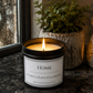 A lit candle in a black glass jar sits on a dark countertop beside a window, with a potted green plant and a decorative lantern in the background. The label reads “HOME” and “IT’S NOT A PLACE IT’S A FEELING.