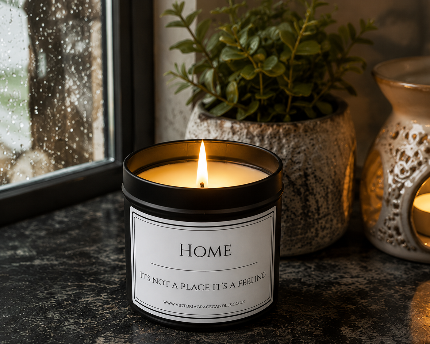 A lit candle in a black glass jar sits on a dark countertop beside a window, with a potted green plant and a decorative lantern in the background. The label reads “HOME” and “IT’S NOT A PLACE IT’S A FEELING.