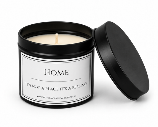 A black metal candle tin with its lid leaning against it, showing a white wax candle and a white label that reads “HOME It’s Not A Place It’s A Feeling.”.