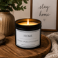 A black glass jar candle with a white label reading 'HOME IT'S NOT A PLACE IT'S A FEELING' sits lit on a round wooden tray, styled with a beige ribbed vase of greenery, a framed 'stay home' sign, and a knitted blanket.