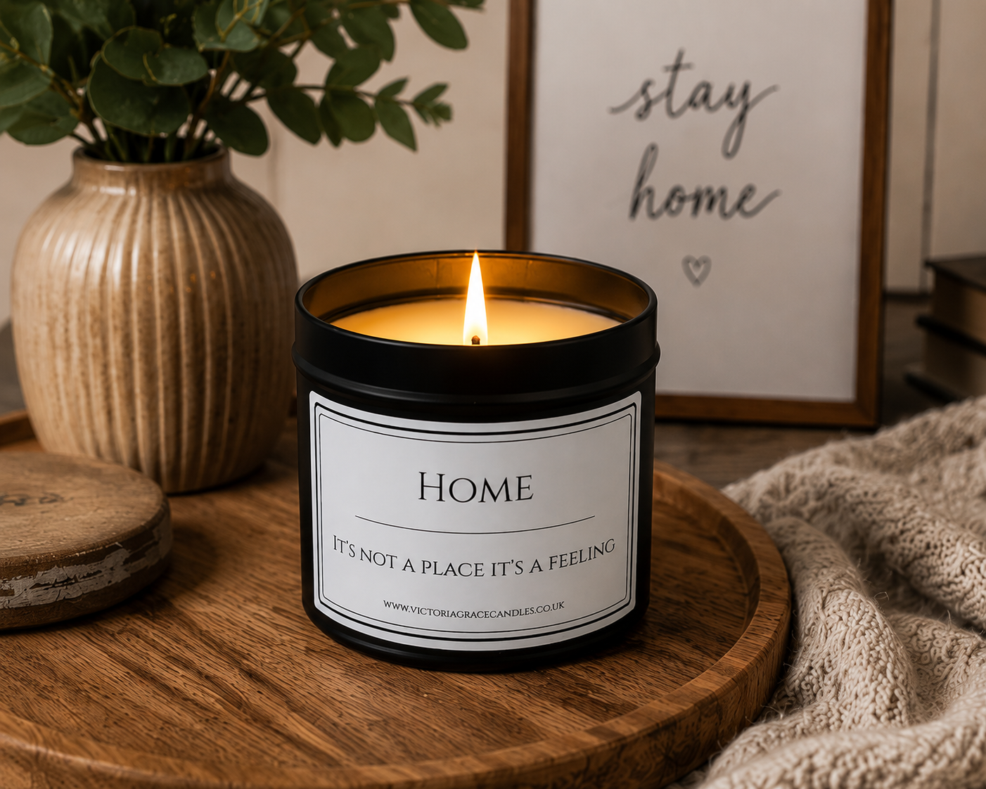 A black glass jar candle with a white label reading 'HOME IT'S NOT A PLACE IT'S A FEELING' sits lit on a round wooden tray, styled with a beige ribbed vase of greenery, a framed 'stay home' sign, and a knitted blanket.