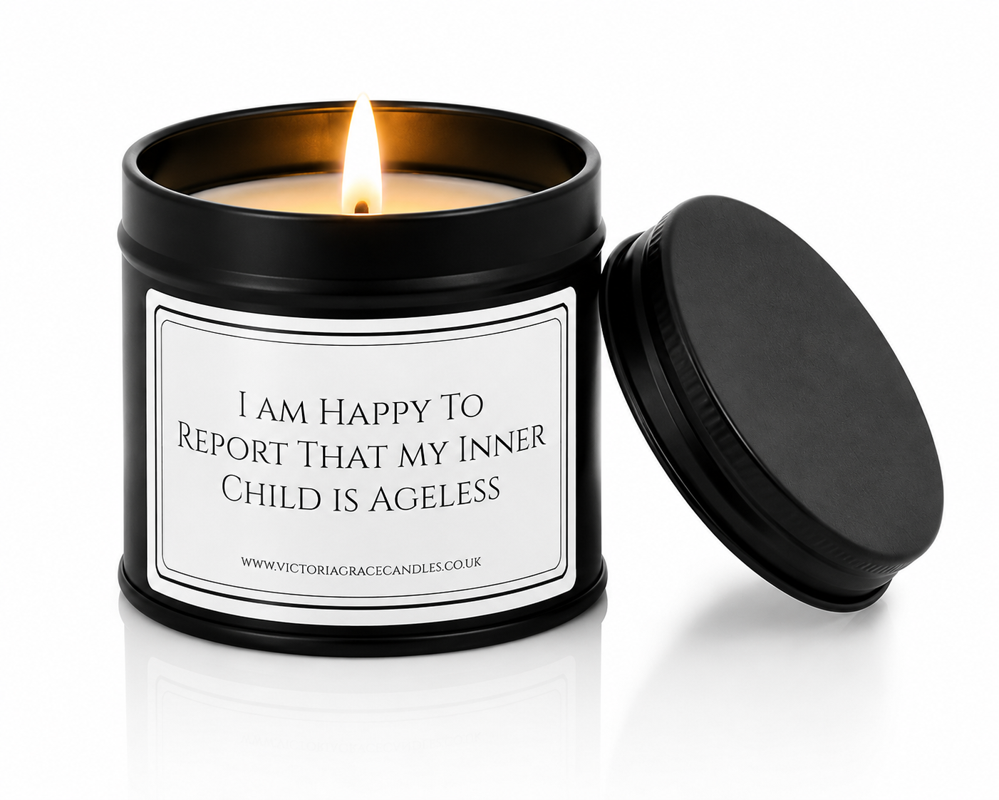 A black metal candle tin with a lit candle and its lid leaning against it on a white background. The front label reads, 'I AM HAPPY TO REPORT THAT MY INNER CHILD IS AGELESS'
