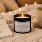 A black candle jar with a white label reading 'I AM HAPPY TO REPORT THAT MY INNER CHILD IS AGELESS' sits on a beige knit blanket next to a book and a ceramic mug, with warm fairy lights blurred in the background.