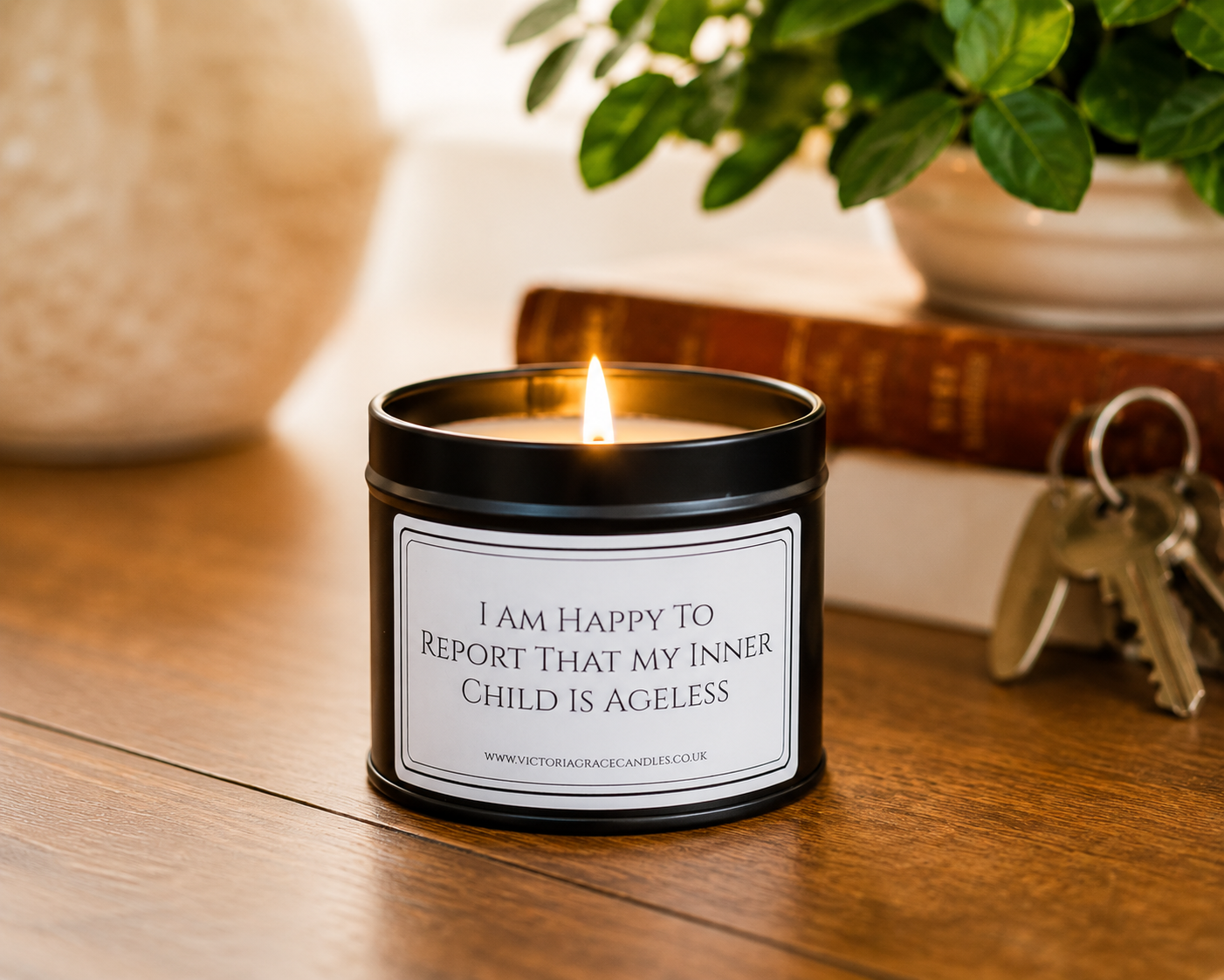 A black metal candle tin with a lit flame sits on a wooden table. The front label reads, 'I AM HAPPY TO REPORT THAT MY INNER CHILD IS AGELESS.