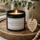 A black metal candle tin with a lit candle sits on a round wooden tray beside a wooden heart-shaped tag that reads 'Stay young at heart.' The scene includes a vase, greenery, and a strand of wooden beads in a softly styled interior setting.