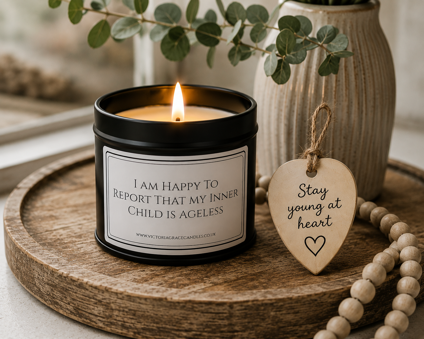 A black metal candle tin with a lit candle sits on a round wooden tray beside a wooden heart-shaped tag that reads 'Stay young at heart.' The scene includes a vase, greenery, and a strand of wooden beads in a softly styled interior setting.