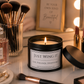 A lit black candle jar with the label 'JUST WING IT' sits on a vanity table surrounded by makeup brushes, a perfume bottle, lipsticks, and a mirror with light bulbs. A framed sign in the background reads 'BE YOUR OWN KIND of Beautiful'.