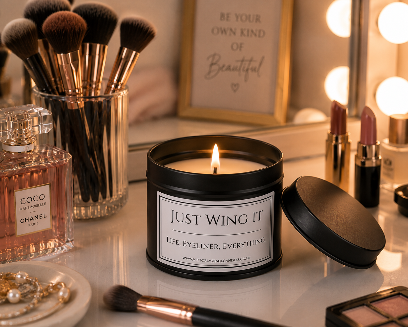 A lit black candle jar with the label 'JUST WING IT' sits on a vanity table surrounded by makeup brushes, a perfume bottle, lipsticks, and a mirror with light bulbs. A framed sign in the background reads 'BE YOUR OWN KIND of Beautiful'.