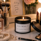 A black metal candle tin with a lit flame sits on a vanity table, surrounded by makeup brushes, lipstick, perfume, jewelry, and an eyeshadow palette. The candle label reads 'JUST WING IT' with the tagline 'LIFE, EYELINER, EVERYTHING'.