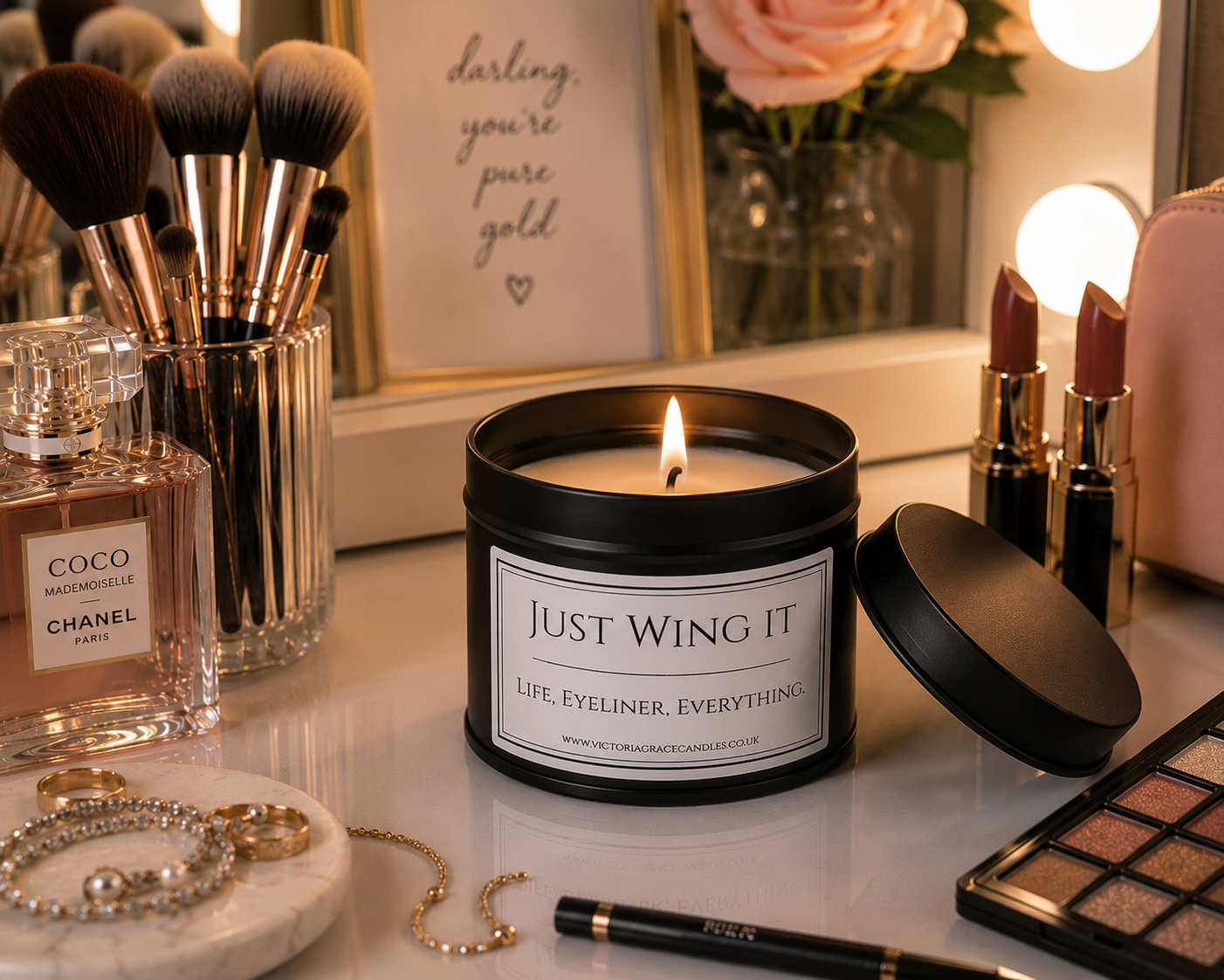 A black metal candle tin with a lit flame sits on a vanity table, surrounded by makeup brushes, lipstick, perfume, jewelry, and an eyeshadow palette. The candle label reads 'JUST WING IT' with the tagline 'LIFE, EYELINER, EVERYTHING'.