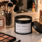 A black metal candle jar labeled 'JUST WING IT' sits on a vanity table with its lid beside it, surrounded by makeup brushes, a Chanel perfume bottle, lipsticks, and an eyeshadow palette in a softly lit mirror setting.