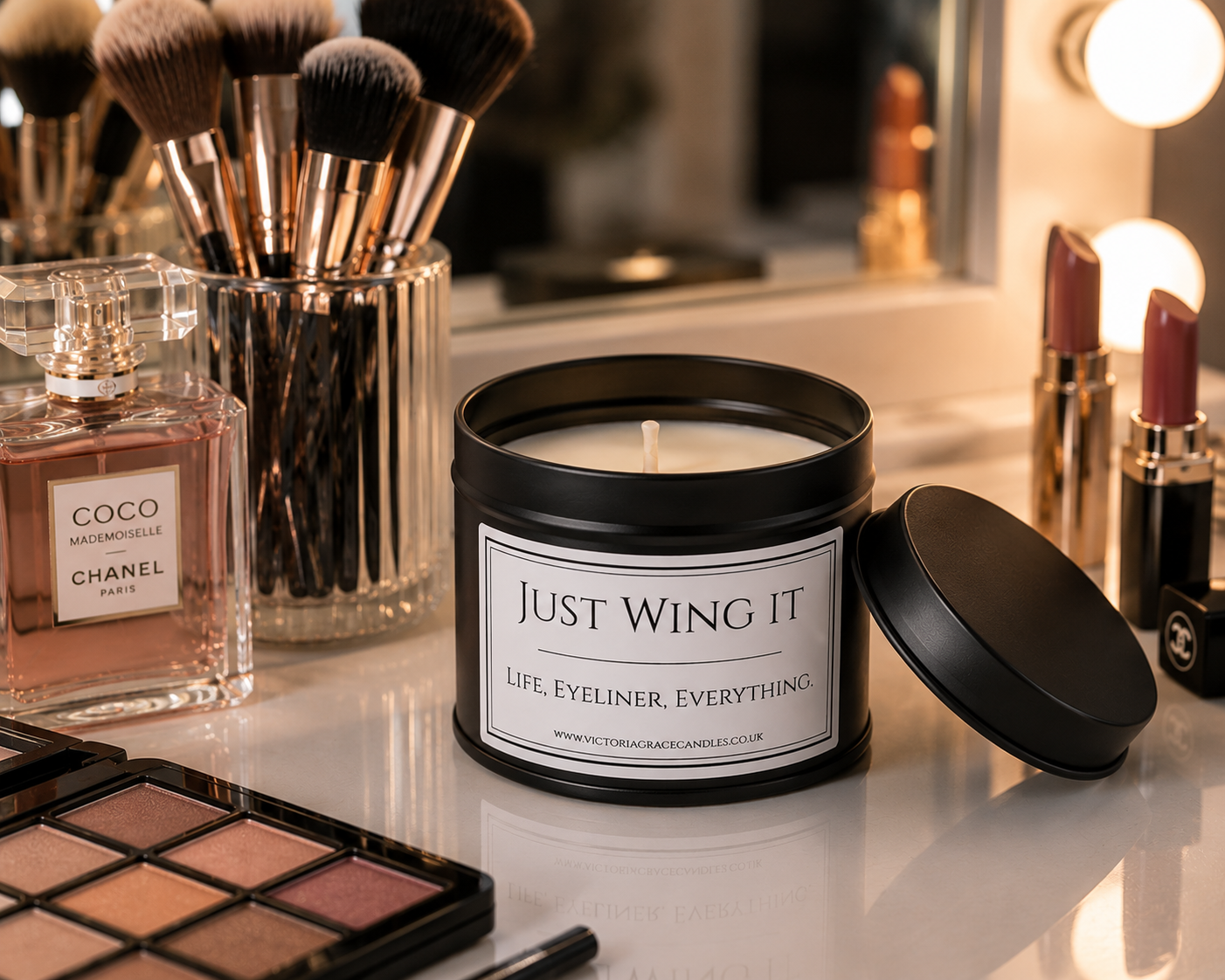 A black metal candle jar labeled 'JUST WING IT' sits on a vanity table with its lid beside it, surrounded by makeup brushes, a Chanel perfume bottle, lipsticks, and an eyeshadow palette in a softly lit mirror setting.