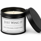 A black metal candle tin with its lid propped open, showing a white wax candle inside. The front label reads 'JUST WING IT' and 'LIFE, EYELINER, EVERYTHING.