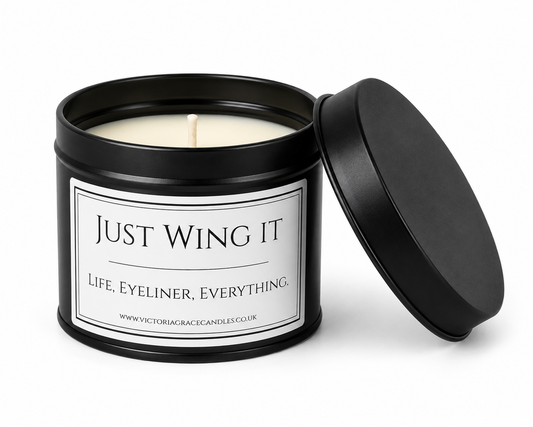 A black metal candle tin with its lid propped open, showing a white wax candle inside. The front label reads 'JUST WING IT' and 'LIFE, EYELINER, EVERYTHING.