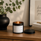 A lit black metal candle tin with a white label sits on a wooden table beside a black vase with eucalyptus and a framed wall print. The label reads, 'LETTING GO IS HARD being free is beautiful