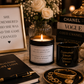 A styled tabletop scene with several black-and-gold self-care and inspirational items: a framed quote print, a lit candle, a black mug with gold text reading 'Empowered Women Empower Woman,' a black pen on a notebook, a stack of Chanel and Vogue books, gold bracelets, and flowers in the background.