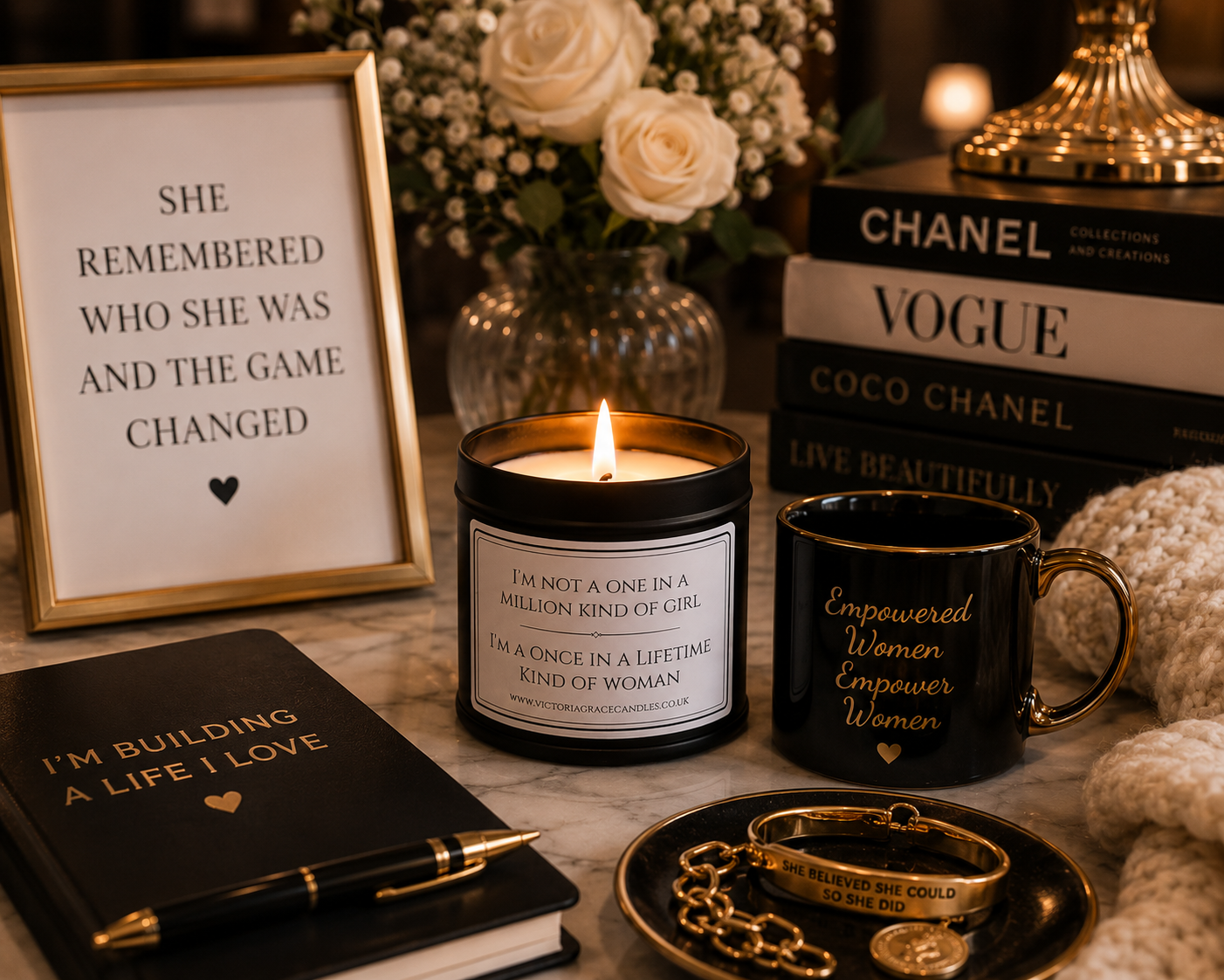 A styled tabletop scene with several black-and-gold self-care and inspirational items: a framed quote print, a lit candle, a black mug with gold text reading 'Empowered Women Empower Woman,' a black pen on a notebook, a stack of Chanel and Vogue books, gold bracelets, and flowers in the background.