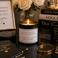 A lit candle in a black jar with a white label reading 'I'M NOT A ONE IN A MILLION KIND OF GIRL' is styled on a dark reflective surface with black mugs, gold jewelry, a framed quote, and books including Chanel and Dior in the background.
