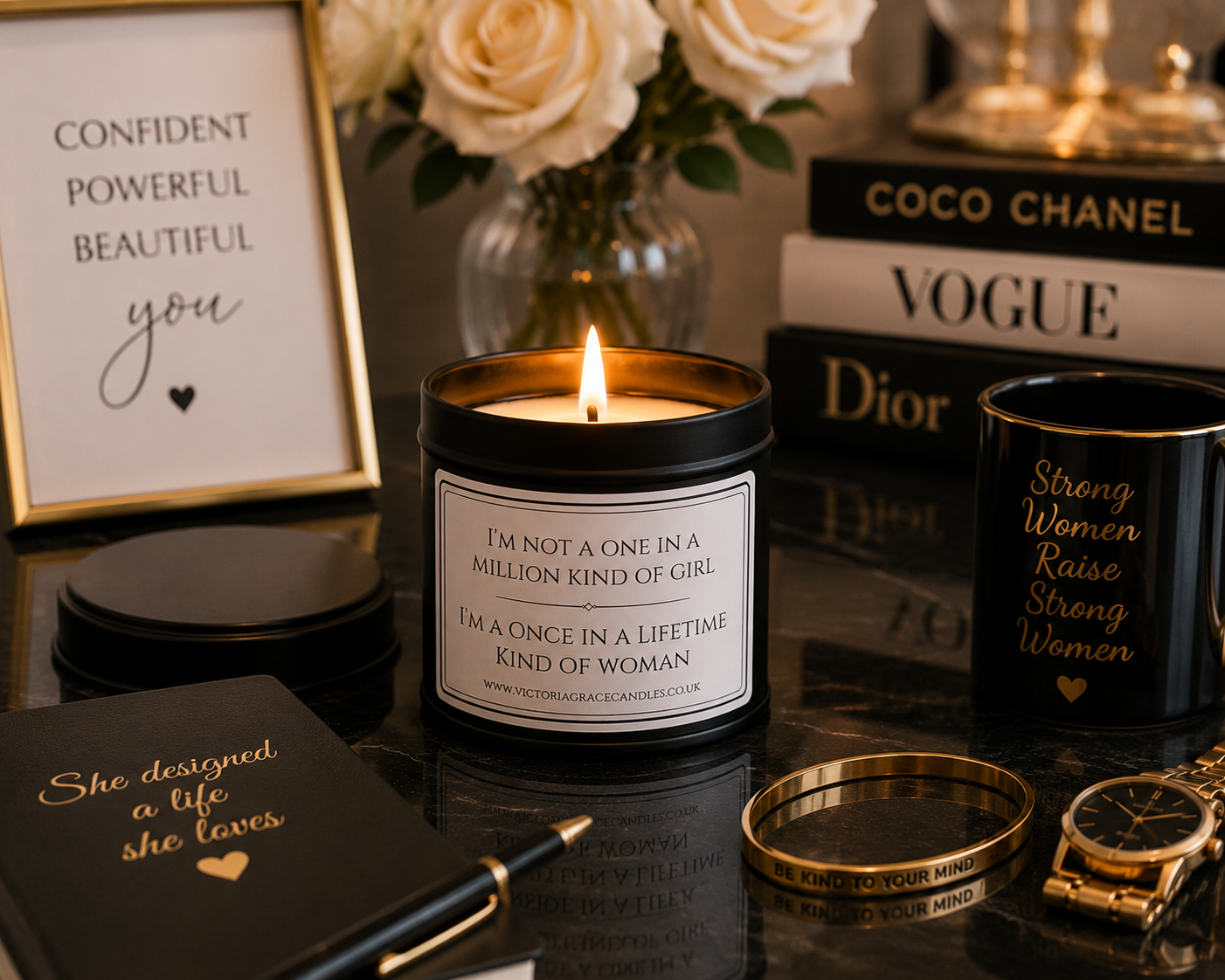 A lit candle in a black jar with a white label reading 'I'M NOT A ONE IN A MILLION KIND OF GIRL' is styled on a dark reflective surface with black mugs, gold jewelry, a framed quote, and books including Chanel and Dior in the background.