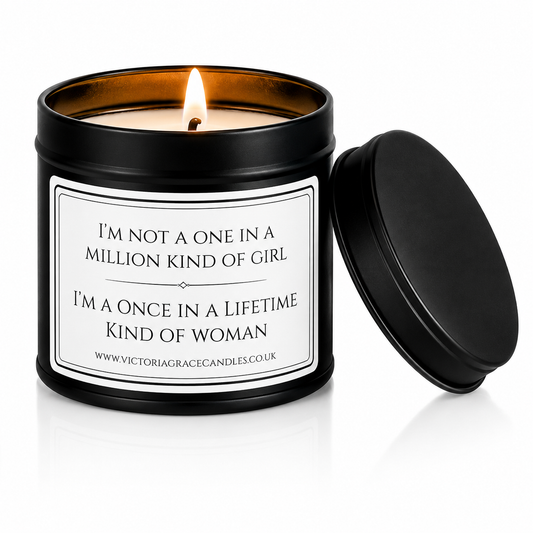 A black metal candle tin with a lit candle inside, shown next to its matching black lid on a white background. The front label reads, 'I'M NOT A ONE IN A MILLION KIND OF GIRL I'M A ONCE IN A LIFETIME KIND OF WOMAN' with the brand Victoria Grace Candles visible at the bottom.