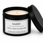 A black metal candle tin with its lid propped open, showing a creamy off-white soy wax candle inside. The front label reads 'SASSY WITH A HINT OF SARCASM' and 'VICTORIA GRACE CANDLES | SOY WAX'.