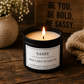 A lit black candle jar labeled 'SASSY' sits on a rustic wooden table, surrounded by a speckled ceramic vase with small white flowers, a knitted beige throw, and a wooden sign in the background.