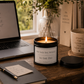 A lit black candle jar with a cream label reading 'SHE SAID SHE WOULD So SHE Did' sits on a wooden desk beside a laptop, notebook, pen, mug, books, and a glass vase with greenery in a bright indoor setting.