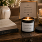 A black candle jar with a lit flame and a matching black lid sits on a rustic wooden table, surrounded by decorative props including a vase of small white flowers, a book, a framed quote, and another candle.