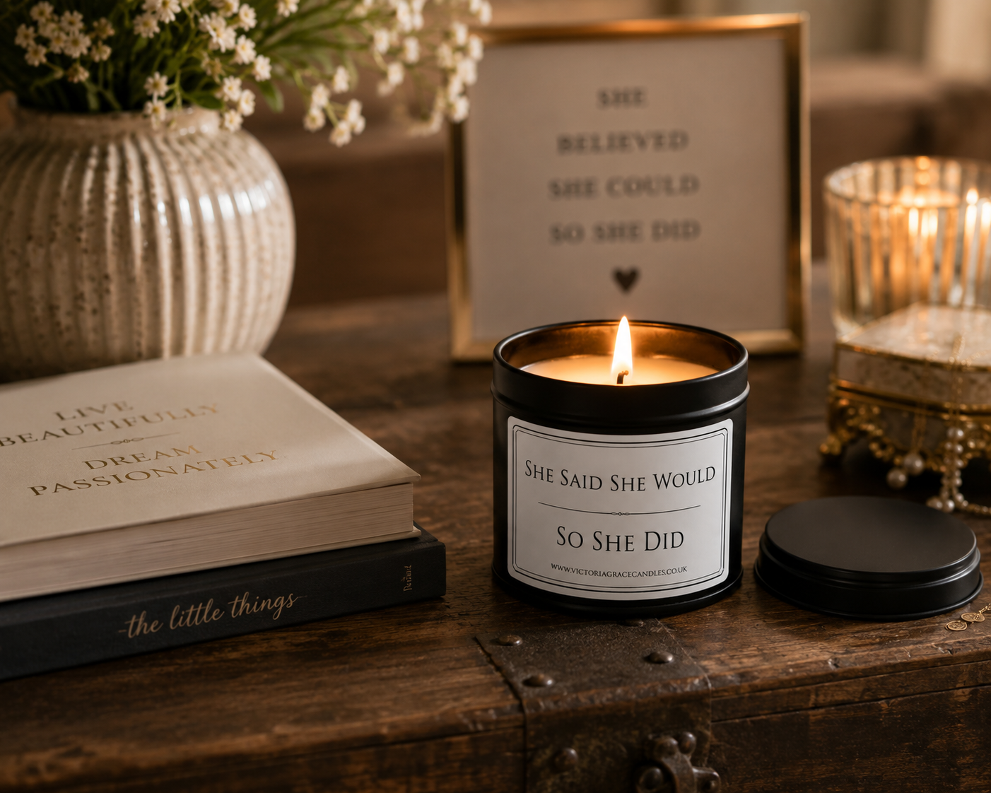 A black candle jar with a lit flame and a matching black lid sits on a rustic wooden table, surrounded by decorative props including a vase of small white flowers, a book, a framed quote, and another candle.