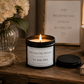 A lit black metal candle jar with a white label reading 'SHE SAID SHE WOULD SO SHE DID' sits on a wooden table, with its lid placed beside it. The scene includes a vase of cream flowers, a framed quote print, a ceramic mug, jewelry, and a book in a styled interior setting.