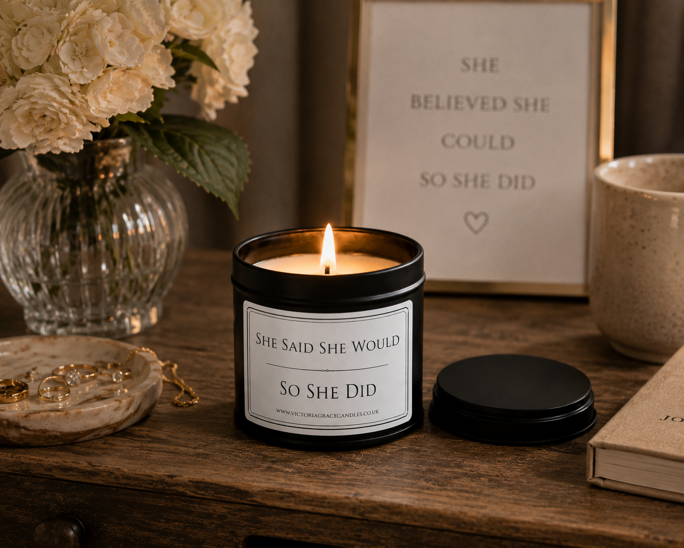 A lit black metal candle jar with a white label reading 'SHE SAID SHE WOULD SO SHE DID' sits on a wooden table, with its lid placed beside it. The scene includes a vase of cream flowers, a framed quote print, a ceramic mug, jewelry, and a book in a styled interior setting.