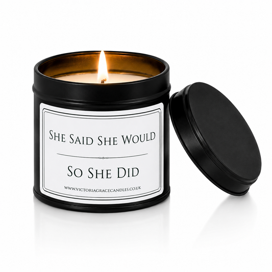 A black metal candle tin with a lit candle inside and its lid leaning against it on a white background. The front label reads 'SHE SAID SHE WOULD SO SHE DID' 