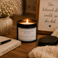 A lit black candle jar with a white label sits on a wooden table beside a closed book, pen, dried flowers in a vase, a knitted blanket, and a framed quote that reads, 'She believed she could, so she did.'.