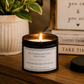 A lit black metal candle tin with a white label sits on a wooden surface in a styled interior setting. A potted plant is blurred in the background, along with stacked books and a sign.