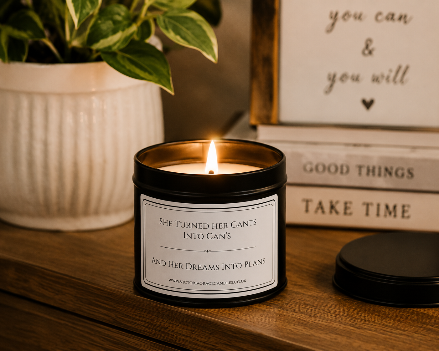 A lit black metal candle tin with a white label sits on a wooden surface in a styled interior setting. A potted plant is blurred in the background, along with stacked books and a sign.