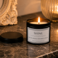 A lit black metal candle jar with a white label reading “SHINE” and “Life is the whole universe is yours,” placed on a dark marble surface with its lid beside it. A warm ribbed glass candle holder and an ornate mirror are blurred in the background.