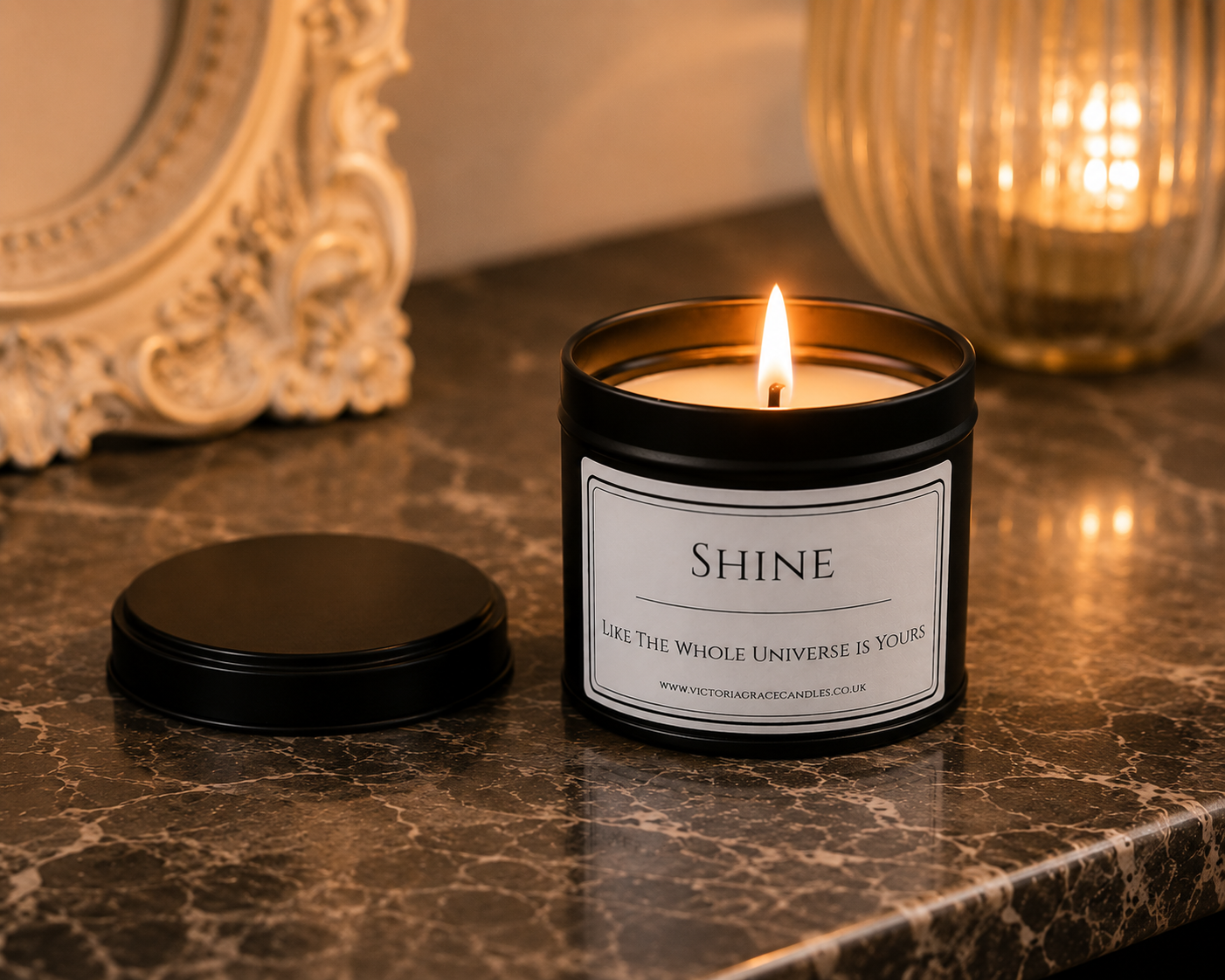 A lit black metal candle jar with a white label reading “SHINE” and “Life is the whole universe is yours,” placed on a dark marble surface with its lid beside it. A warm ribbed glass candle holder and an ornate mirror are blurred in the background.
