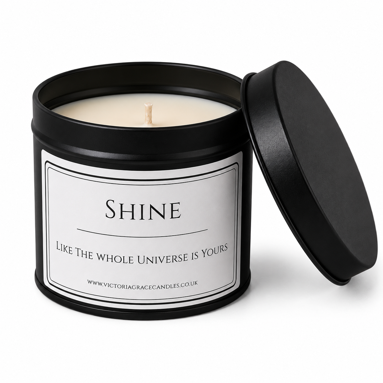 A black metal candle tin with its lid propped open, showing a cream-colored wax candle inside. The front label is white with black text reading 'SHINE' and 'LIKE THE WHOLE UNIVERSE IS YOURS', with the brand Victoria Grace Candles visible at the bottom.