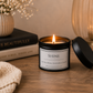 A lit candle in a black metal tin with a white label reading “SHINE,” placed on a wooden table with its lid beside it. The scene includes stacked books, a textured cream towel, a vase with dried flowers, and a warm glowing glass vessel in the background.