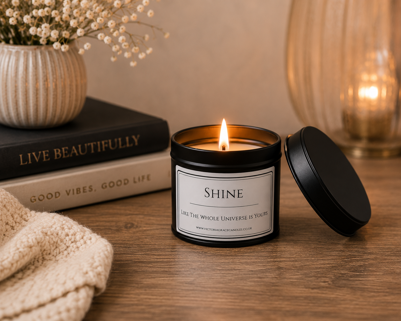 A lit candle in a black metal tin with a white label reading “SHINE,” placed on a wooden table with its lid beside it. The scene includes stacked books, a textured cream towel, a vase with dried flowers, and a warm glowing glass vessel in the background.