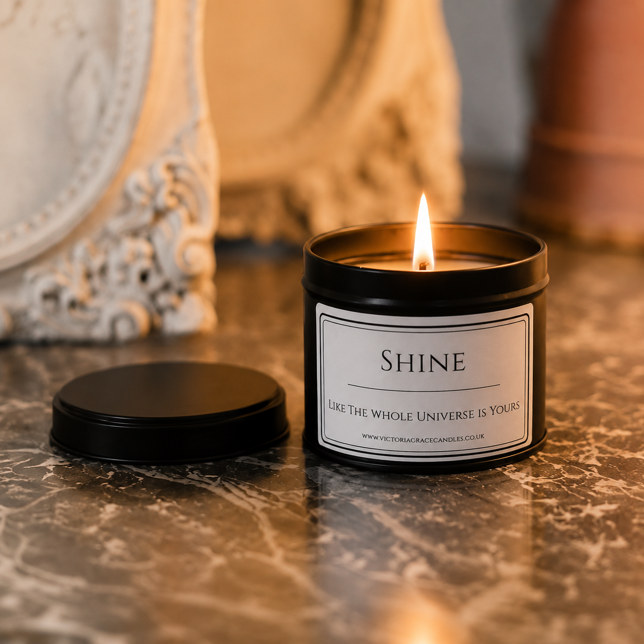 A black metal candle jar with a white label reading 'SHINE' and a visible flame, shown on a marble surface with its lid placed beside it. The background includes warm-toned decorative objects in a softly lit indoor setting.