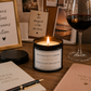 A lit candle in a black jar labeled 'The Naughty Corner' sits on a wooden desk surrounded by romantic and motivational props, including a glass of red wine, notebooks, framed quotes, and a vase.