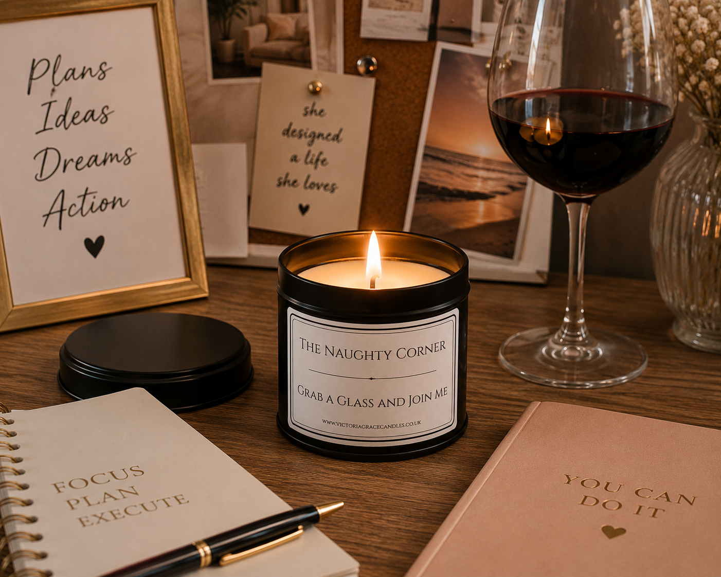 A lit candle in a black jar labeled 'The Naughty Corner' sits on a wooden desk surrounded by romantic and motivational props, including a glass of red wine, notebooks, framed quotes, and a vase.