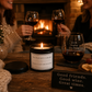 A cozy living room scene with two women holding wine glasses near a fireplace, with a candle and a wine glass engraved with quotes on a wooden table in the foreground.