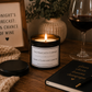A lit candle in a black jar labeled 'The Naughty Corner' sits on a wooden table with a wine glass, a knit blanket, a notebook, and a framed sign in a cozy indoor setting.