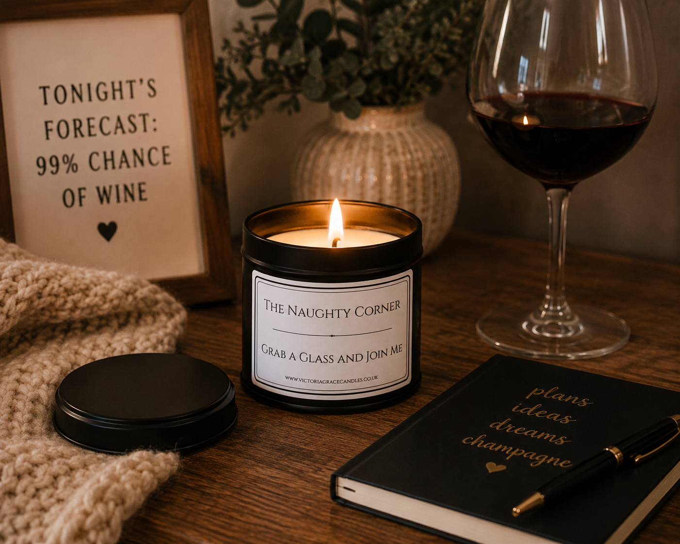 A lit candle in a black jar labeled 'The Naughty Corner' sits on a wooden table with a wine glass, a knit blanket, a notebook, and a framed sign in a cozy indoor setting.