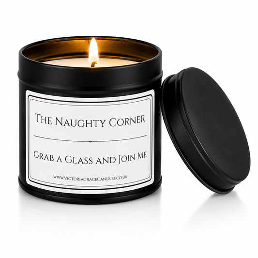 A black metal candle tin with a white label reading 'The Naughty Corner' and 'Grab a Glass and Join Me,' with the flame lit and the matching lid leaning against it on a white background.