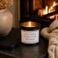 Luxury motivational candle beside a glowing fireplace with a cosy knitted blanket and soft candlelight, creating a warm and relaxing setting.