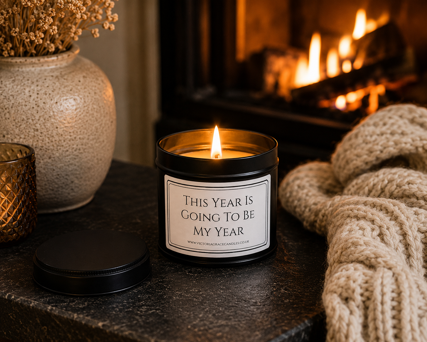 Luxury motivational candle beside a glowing fireplace with a cosy knitted blanket and soft candlelight, creating a warm and relaxing setting.