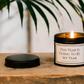 Luxury inspirational soy wax candle displayed on a rustic wooden table beneath tropical green leaves, creating a calm and motivating atmosphere.